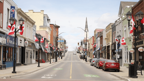 Newmarket, ON. A small-town street lined with historic buildings, adorned with Canadian flags. Cars are parked along the roadside, conveying a peaceful, patriotic scene.