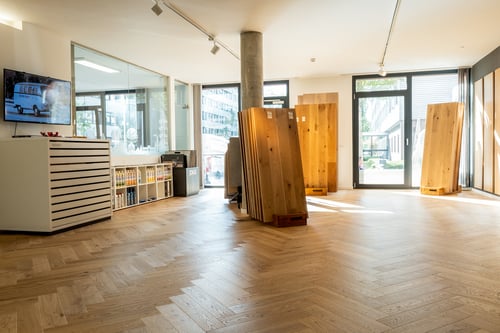 Moderner Showroom mit Fischgrät-Parkett, großen Holzmustern, Pflegeprodukten und viel Tageslicht durch große Fenster.