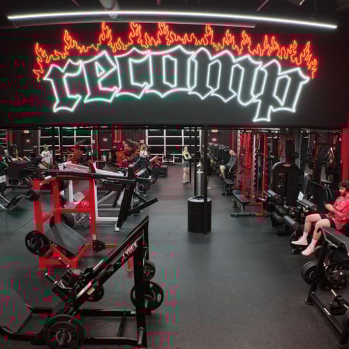 North Richland Hills gym with strength training machines, free weights & members exercising under glowing neon “recomp” sign.