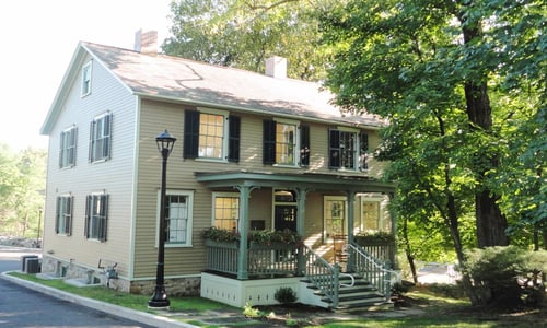 Beige colonial-style building with black shutters and green porch housing William Pitt Sotheby's International Realty in North Stamford, Connecticut, surrounded by mature trees.