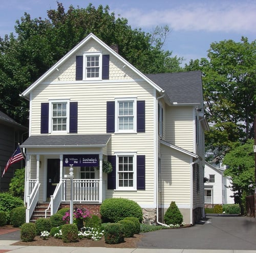 Yellow colonial-style building with navy shutters and front porch housing William Pitt Sotheby's International Realty in Darien, Connecticut, featuring American flag and landscaped foundation plantings