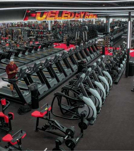 Spacious Lewisville gym with rows of treadmills, ellipticals, and strength machines under bright lighting and a “Fueled by Faith” sign reflected in wall mirrors.