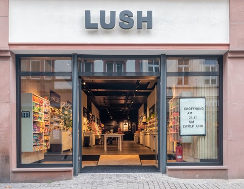 Exterior view of shop. Large entrance in centre with Lush sign above. Illuminated cinema style lightbox display in the right window. Full interior of shop can be seen from the outside.
