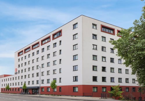 Sechsgeschossiges, modernes Gebäude mit weißer Fassade und rotem Sockel, vielen gleichmäßigen Fenstern und roten Rahmen im oberen Stockwerk; davor verläuft eine breite Straße mit wenigen Bäumen und Fahrrädern am Eingangsbereich unter blauem Himmel.