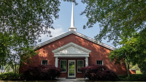 The Church of Jesus Christ of Latter-day Saints meetinghouse in Dayton, Tennessee. The viewer is presented with a red brick building, pitched roof, and one white steeple. The front door entryway is protected by a white pitched roof supported by two white columns. Bushes and two large trees form the front landscaping.