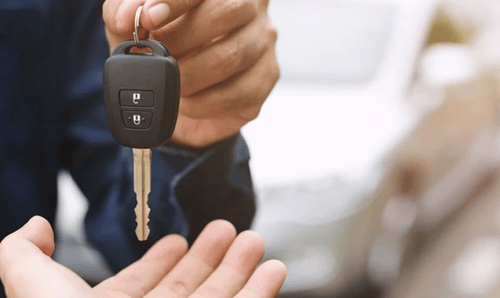 A person holds out a car key with buttons, preparing to hand it to another person whose hand is open to receive it.