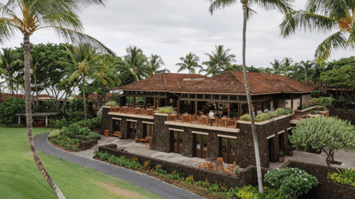 Tropical restaurant with stone walls, wooden balconies and an open-air dining area, surrounded by lush greenery and palm trees