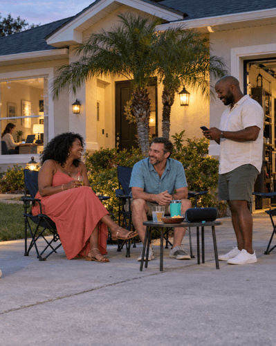 Family and friends gather in an Orlando driveway at dusk as children draw with chalk, an adult uses a phone beside a speaker, and someone works on a laptop inside.