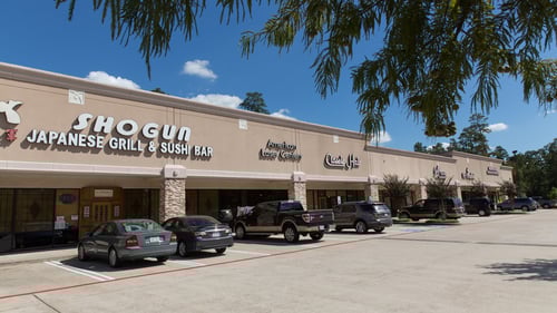 Cars parked along entrance to small shops at Windvale Shopping center with a tree hanging over the parking lot