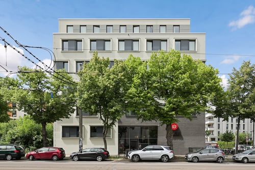 Modernes mehrstöckiges Gebäude mit heller Fassade und großen Fenstern, im Erdgeschoss ein Eingang mit Glasfront, davor eine Reihe geparkter Autos und hohe Bäume, deren Kronen einen Teil der Fassade verdecken; darüber verlaufen Oberleitungen vor blauem Himmel mit wenigen Wolken