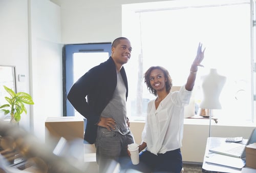 Two people in a bright space discuss ideas. A woman, smiling, gestures upward with enthusiasm, holding a coffee cup. A man stands beside her, listening intently.