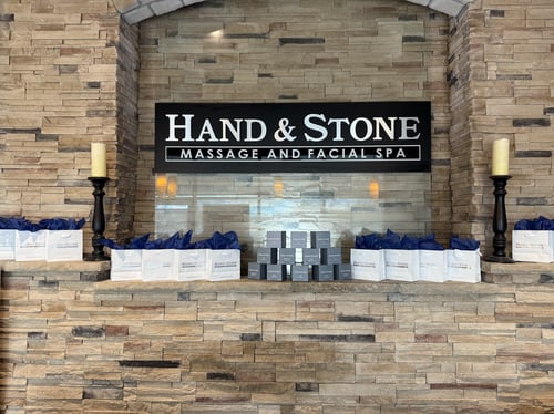 Hand & Stone Massage and Facial Spa sign mounted above a stone fireplace mantel. The mantel is decorated with symmetrical arrangements of white gift bags with blue tissue paper, a stack of small retail product boxes, and two tall black candlesticks.