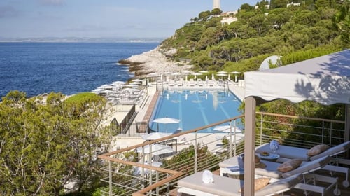 Club Dauphin overlooking the sea, featuring an outdoor swimming pool, lounge chairs, shaded seating areas, lush greenery and a hill in the background