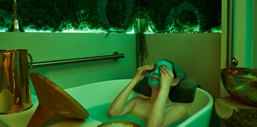 Person sits in a white bath in a green lit room. They have product covering their face and is placing eyemasks onto their eyes. A shelf lays cross the bathtub with wooden mirror and a brass water jug. To the right of the bathtub is a wooden pedestal with a brass bowl.