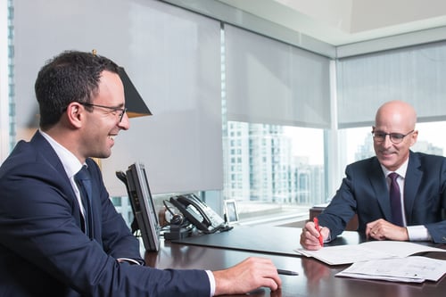 Two male professionals in navy suits sitting across from each other at a dark wooden desk in a modern office with floor-to-ceiling windows overlooking a city skyline. They appear to be in a meeting or consultation, with documents on the desk between them. The office features contemporary furnishings including a desk lamp and office equipment, creating a polished, corporate atmosphere.