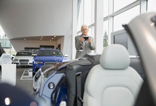 An elderly man smiles while taking a photo with his smartphone inside a bright car showroom. He stands near a blue car, with others displayed around.