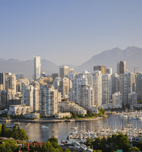 Vancouver skyline with tall, modern buildings reflecting in calm water under a clear blue sky, mountains in the backdrop, and boats docked at a marina.