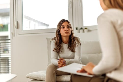 Woman seated on a couch talking with a counselor holding a clipboard in a therapy session.
