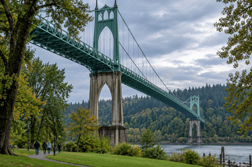 St. Johns Bridge rises above Cathedral Park in Portland, Oregon, with visitors walking beneath leafy trees beside the Willamette River under a cloudy sky.