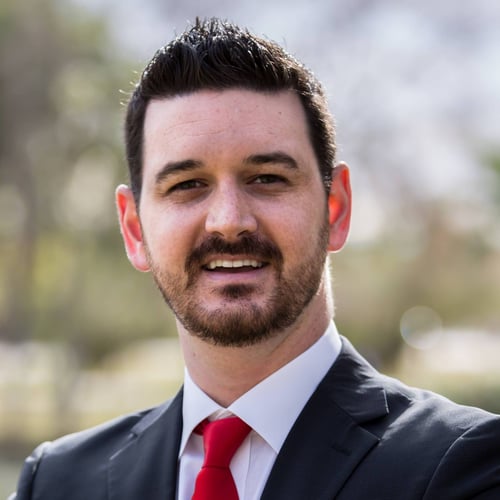 Agent Ryan McBride standing and smiling wearing white button up shirt with red tie and black blazer