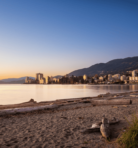 A tranquil beach at sunset with scattered driftwood logs, calm water reflecting the orange and blue sky, and a cityscape backed by forested hills.