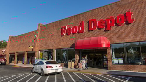 White car passing two people with shopping car exiting Food Depot with red awning at Sweetwater village