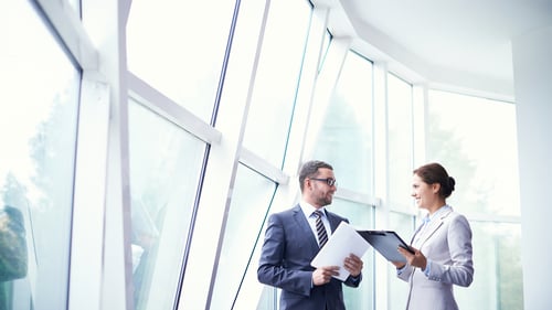 Professionals in business attire engage in a discussion, holding documents, against a backdrop of large, modern windows.