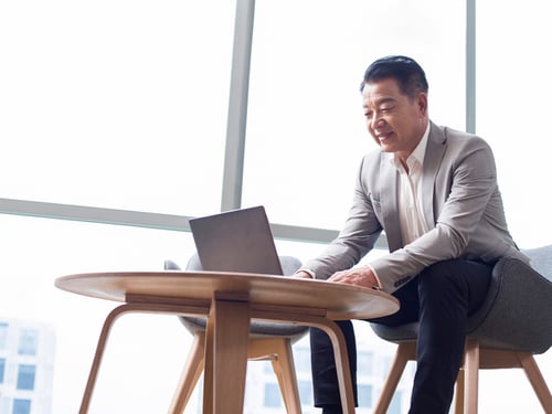 A man in a gray suit sits on a gray chair, smiling and using a laptop on a wooden table. Bright, modern office with large windows in the background.