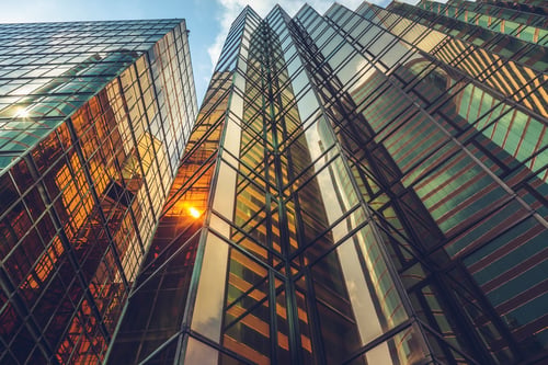 Upward view of modern glass office towers with reflective facades and sunlight shining between the buildings.
