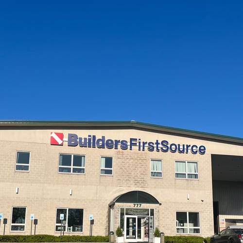 Front exterior of Builders FirstSource building with large company signage, beige brick facade, multiple windows, and a designer showroom entrance with glass doors, set against a clear blue sky and parking lot.