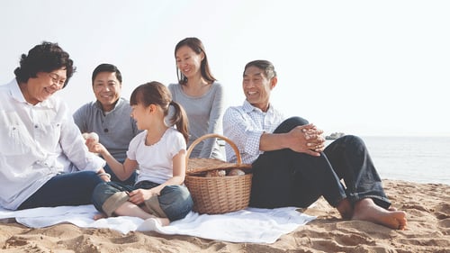 A joyful family sits on a beach enjoying a picnic. They share smiles under a bright sky. A wicker basket sits on a blanket, adding to the warm, relaxed atmosphere.