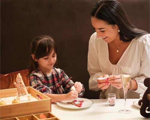Smiling woman and young girl decorating holiday cookies together at a table, with festive decorations and glasses of sparkling drink.