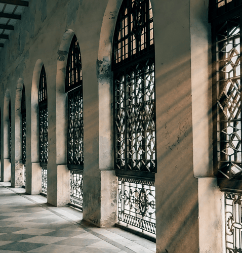 Sunlight streams through ornate, arched windows casting intricate shadows on a stone corridor. The atmosphere is serene and historic.