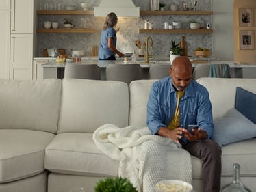 Man sitting on a couch looking at the Kidde App while woman is cooking in the background