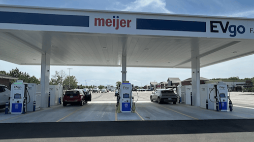 Several EVgo fast charging units installed in marked parking spaces at a public charging station, with EVgo logos on the chargers, protective bollards around each unit. NACS and CCS chargers featured in the image. Protective canopy covers the station.,