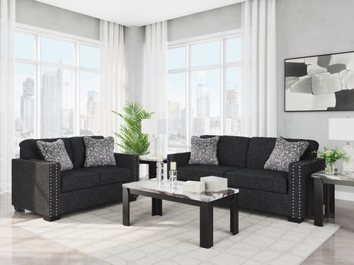 Modern living room with two dark sofas, patterned pillows, a coffee table, side tables with lamps, abstract art, large windows, and a cityscape view in the background.