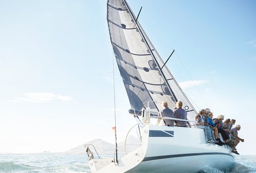 A group of people sail on a yacht in clear, sunny weather. The sail is fully extended, and the ocean is calm. There’s land in the distant background.