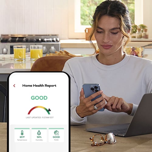 A woman checks her phone, in the foreground a phone with a screen showing the indoor air quality is good.