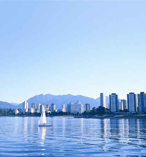 A white sailboat on calm water, with a city skyline and tall buildings in the background. Mountains and clear blue sky create a serene setting.