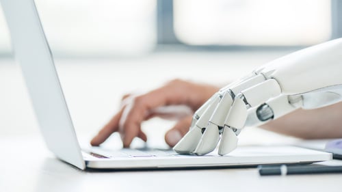 A human and a robotic hand type on a laptop, symbolizing collaboration between humans and technology. The setting is bright, suggesting innovation.