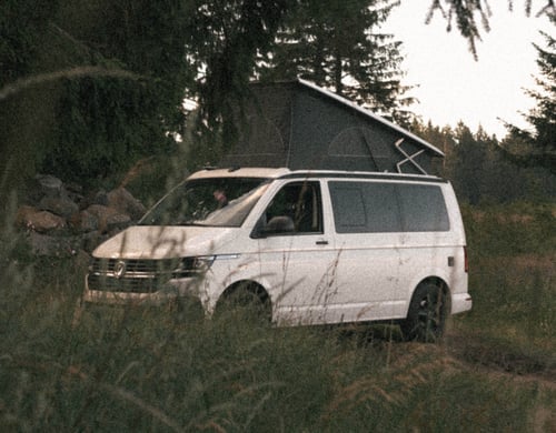 Weißer VW California Beach Camper mit aufgestelltem Dach auf einer Waldlichtung