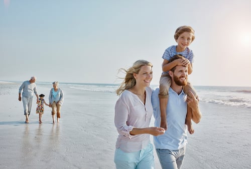 A family enjoys a sunny day on the beach. A man carries a smiling child on his shoulders, walking alongside a woman. An elderly couple and child stroll behind them.