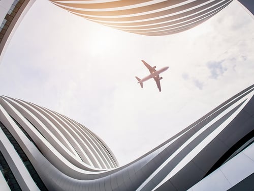 A plane flies above a futuristic building with curved, wavy lines and a bright sky. The perspective creates a sense of openness and modernity.