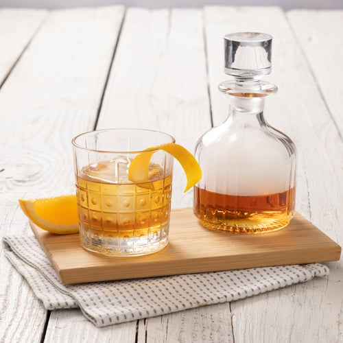 Old fashioned cocktail in a patterned glass with an orange peel garnish, served beside a glass decanter of whiskey