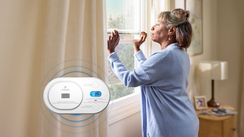 A woman opens her window. In the foreground, a carbon monoxide alarm is going off, showing that 39 ppm has been detected in the air.  Blue circles representing the sound is emanating from the device.