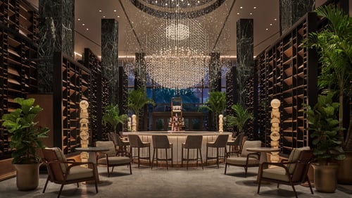 Luxurious hotel bar with a grand chandelier, marble columns, elegant seating, and shelves filled with decor. Large potted plants add a touch of greenery to the space.