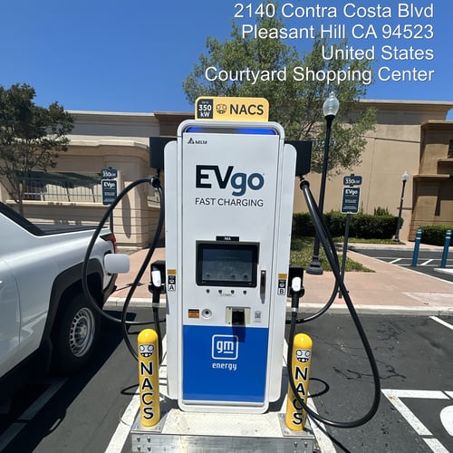 NACS EVgo fast charging unit installed in marked parking space at a public charging station, with EVgo logos on the charger,  and yellow protective bollards around the unit. Close up view.