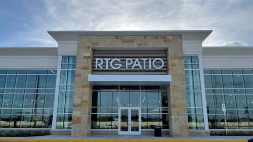 Exterior view of RTG Patio at Rooms To Go entrance in Pearland, TX, featuring modern stone and glass architecture under a bright blue sky.