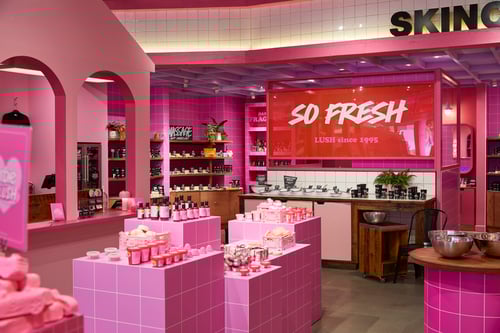 Interior view of shop. Entirely pink themed interior, with the walls and furniture painted in different pink tones. Various packaged and unpackaged products are displayed on units along the walls and on some pedestal units in the middle of the floor. A large sign on the backwall reads So Fresh Lush Since 1995.