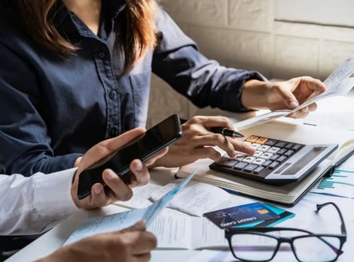 Two persons calculating their finances using a phone and a calculator. Bills, glasses and a credit card are on the table.